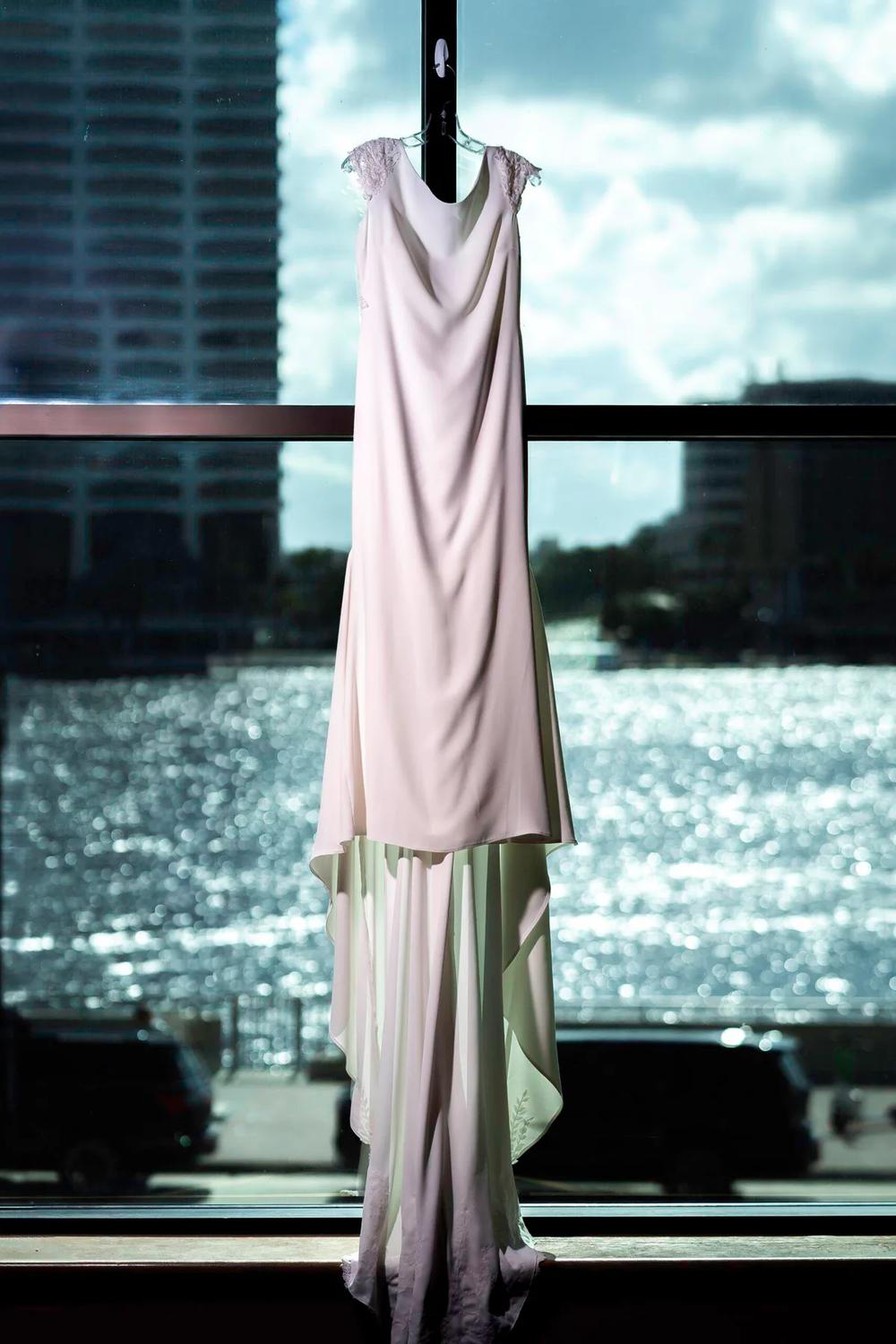 A delicate wedding dress hangs elegantly against a shimmering waterfront backdrop under soft natural light.