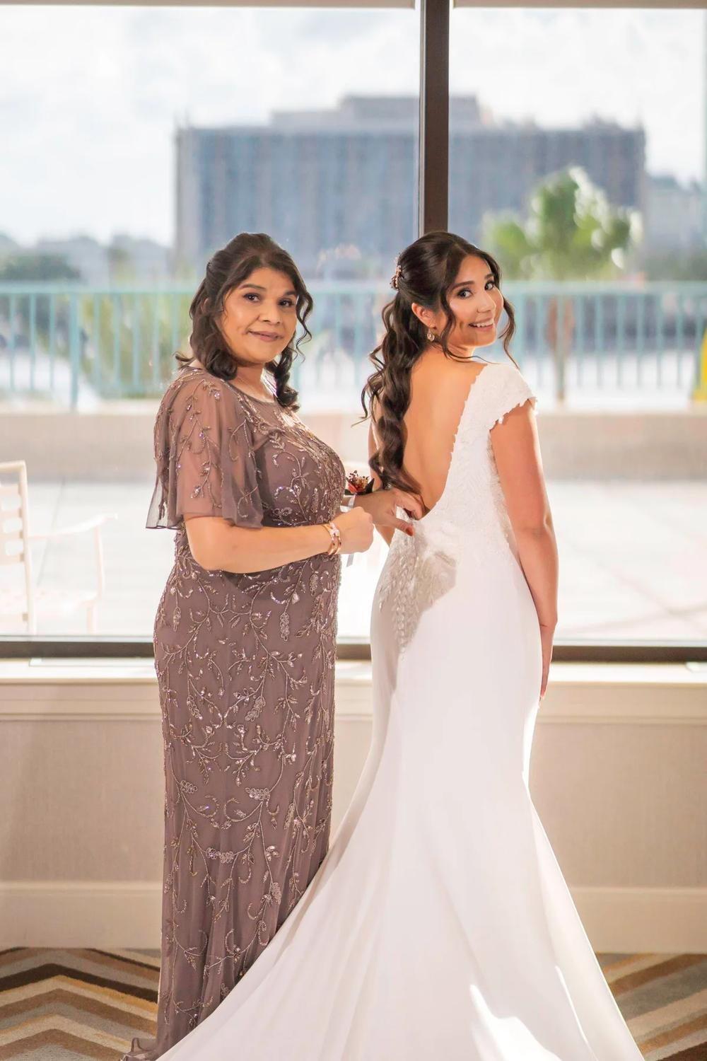 A bride and her mother share a joyful moment in front of a large window with an urban view.