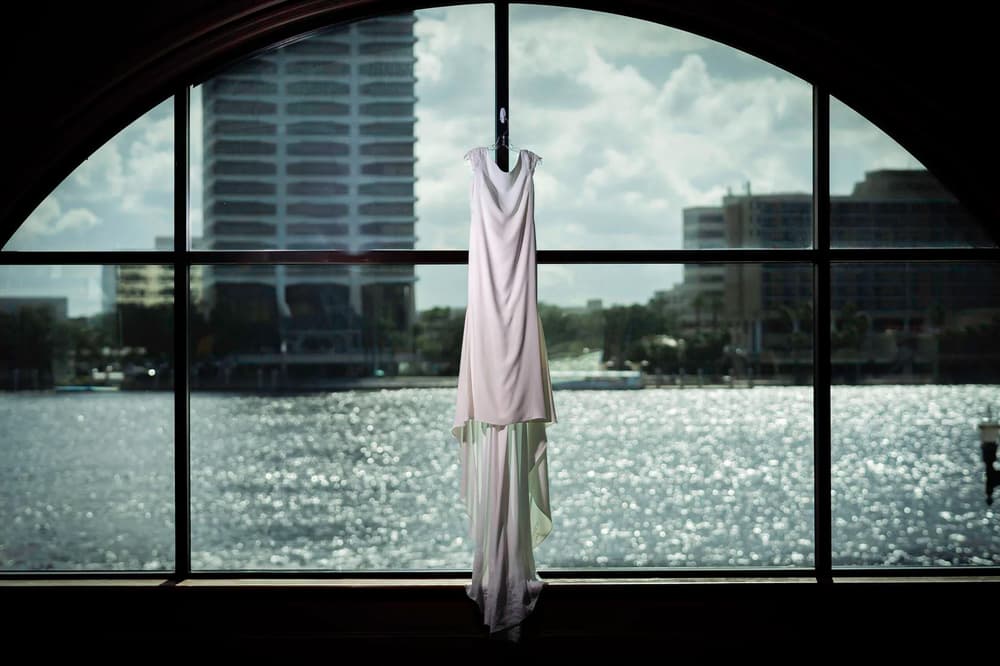 A wedding dress hangs elegantly against a large window overlooking a sparkling waterfront and urban skyline under an overcast sky.