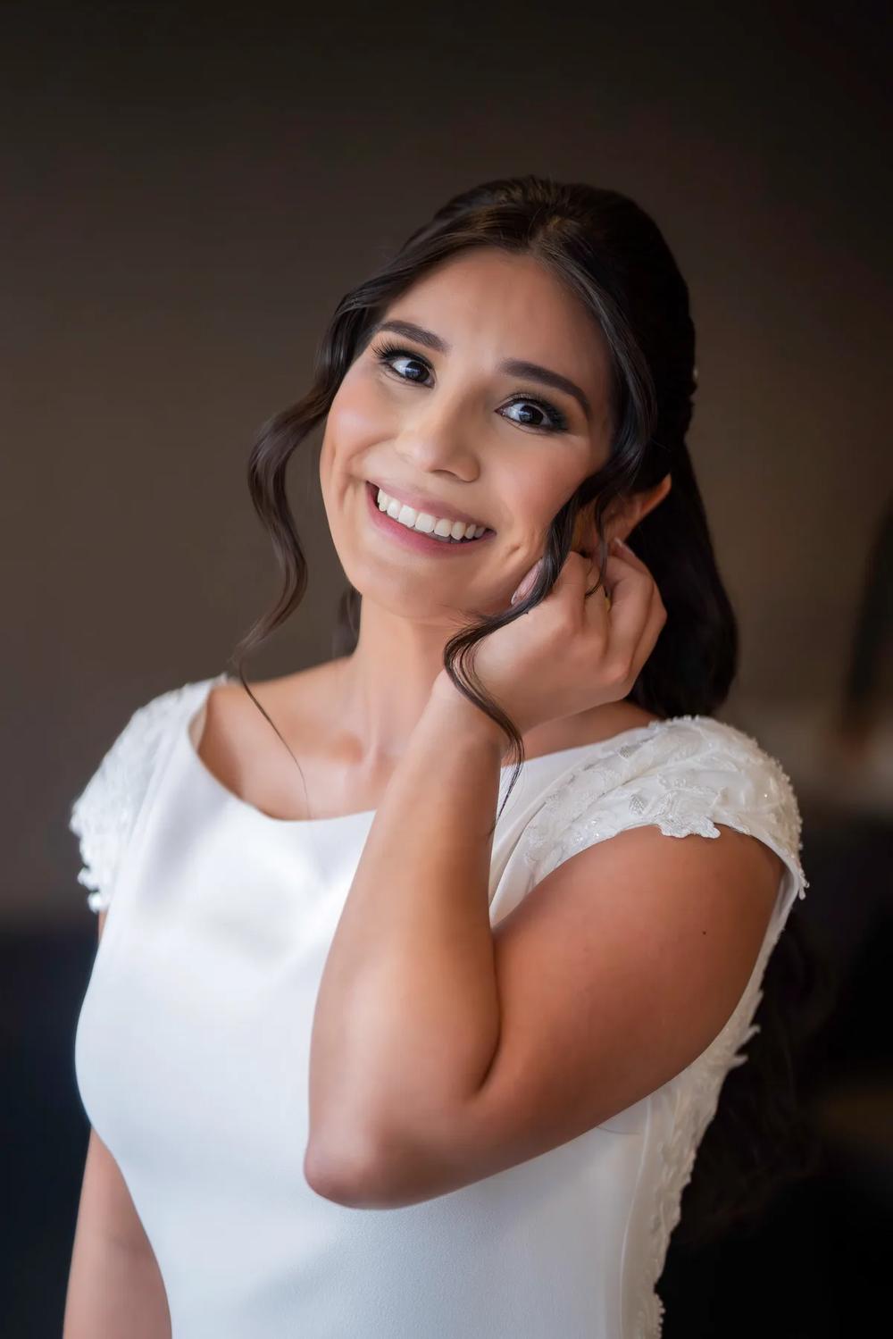 A radiant bride in a white gown smiles warmly as she adjusts her hair against a soft, neutral backdrop.
