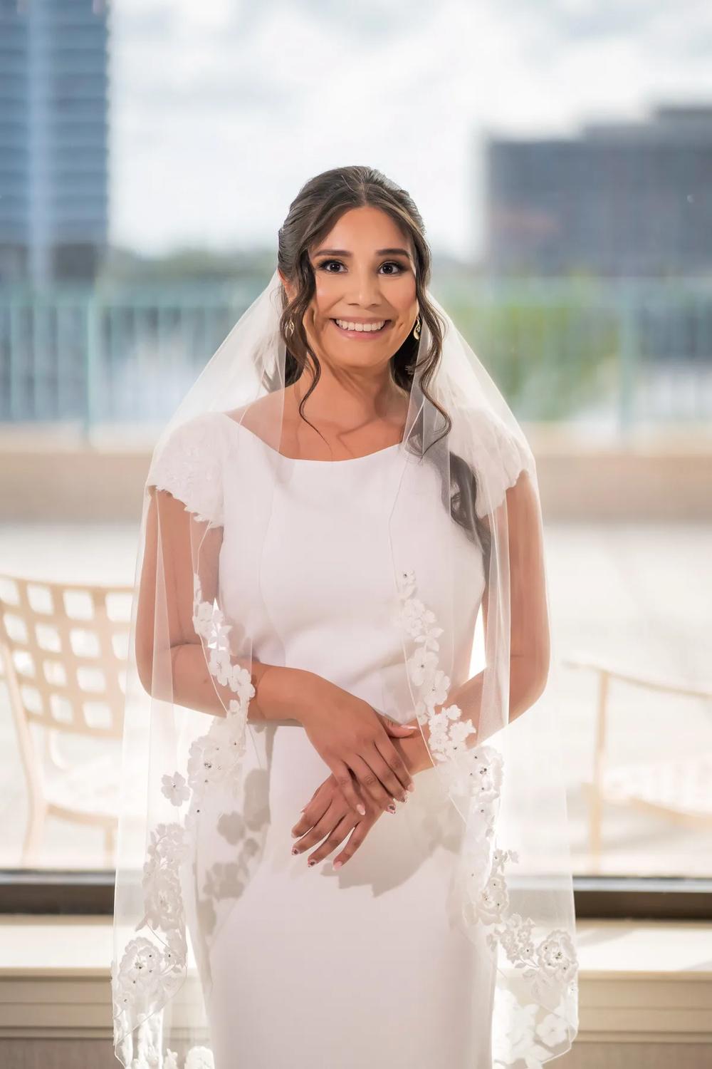A bride in a modern wedding dress and veil smiles brightly against an urban backdrop with soft natural light.
