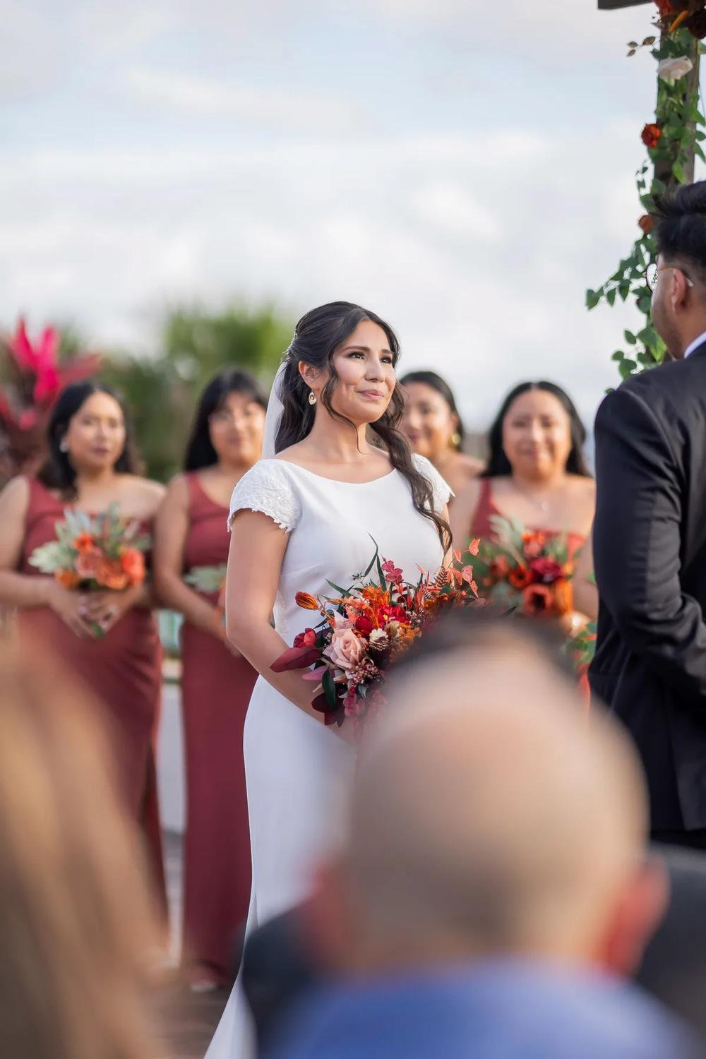 A bride stands at the altar during a wedding ceremony with bridesmaids in burgundy dresses and autumnal bouquets, under soft golden hour lighting.