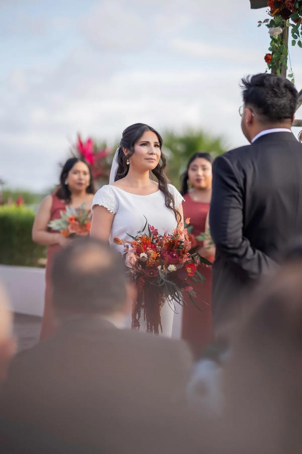 A bride stands at the altar during a wedding ceremony under a cloudy sky, holding her bouquet with an emotional expression.
