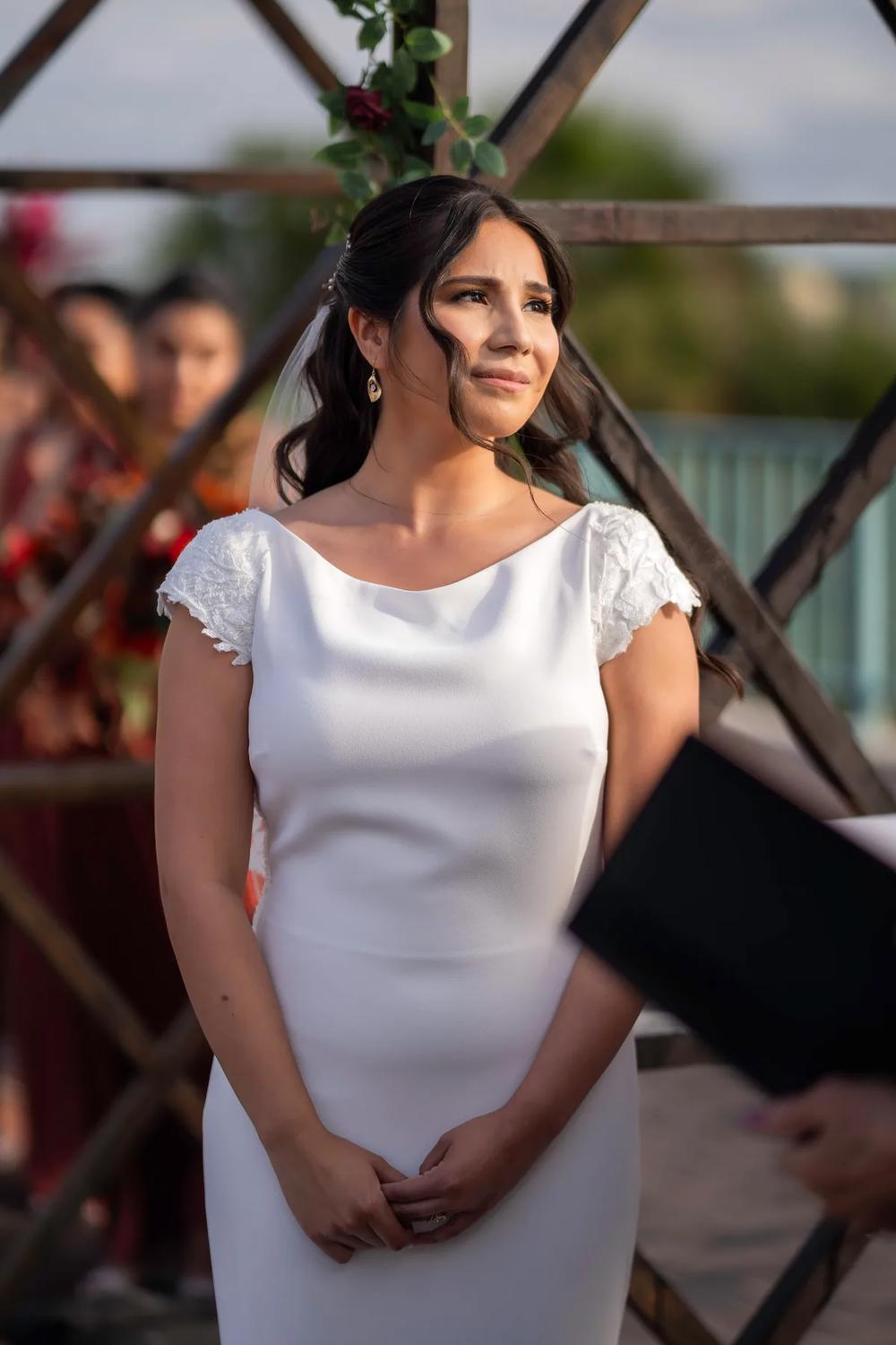 A bride in a white dress stands at an outdoor wedding ceremony with soft golden hour lighting and a serene mood.
