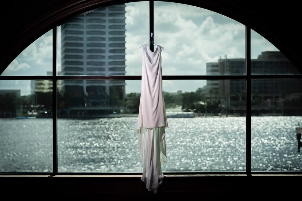 A wedding dress hangs elegantly against a large window overlooking a sparkling waterfront with modern buildings in the background.