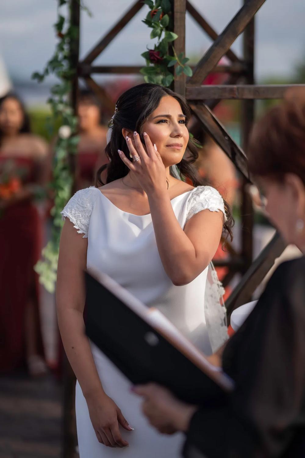 A bride in a white wedding dress adjusts her hair during an outdoor ceremony under golden hour lighting.
