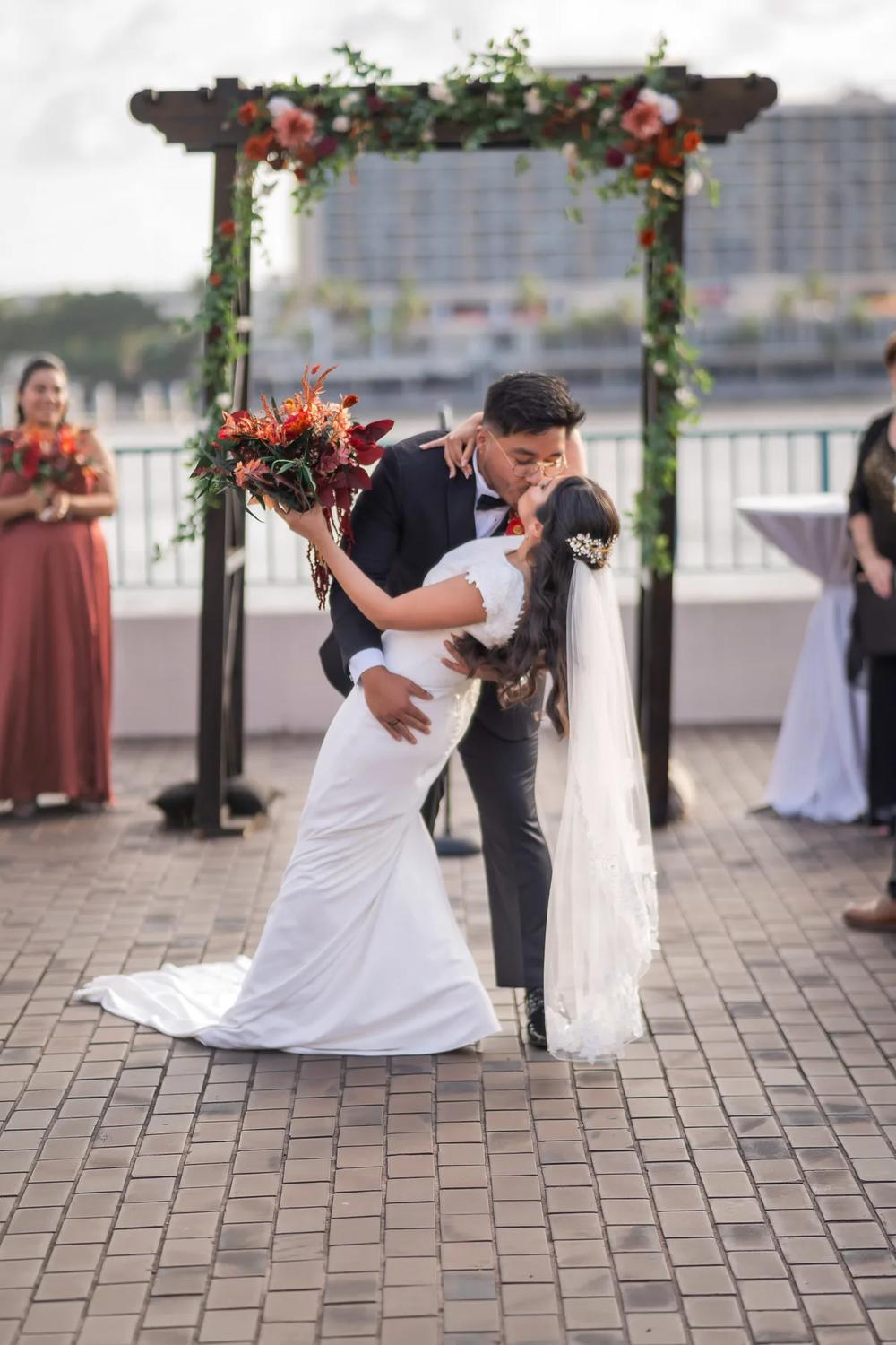 A bride and groom share their first kiss under a floral archway during an outdoor wedding ceremony.