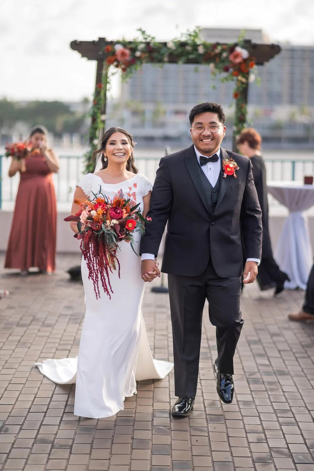 A bride and groom walk hand-in-hand down a brick path under an elegant floral archway during the golden hour.