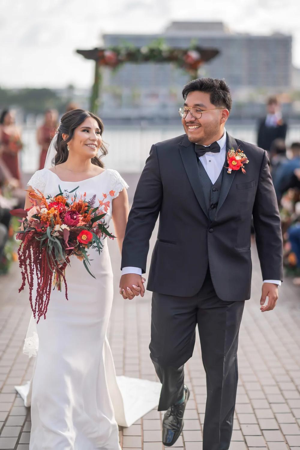 A bride and groom share a joyful moment walking hand-in-hand down an aisle adorned with vibrant autumnal flowers under a clear sky.