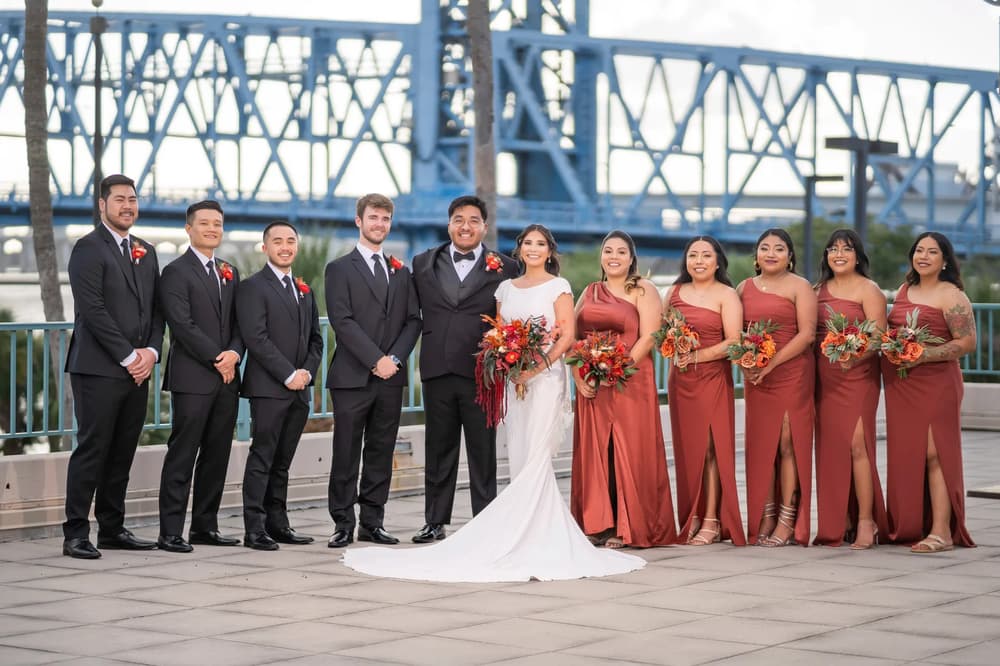A vibrant bridal party poses together on a bridge with a striking blue steel structure in the background under golden hour lighting.