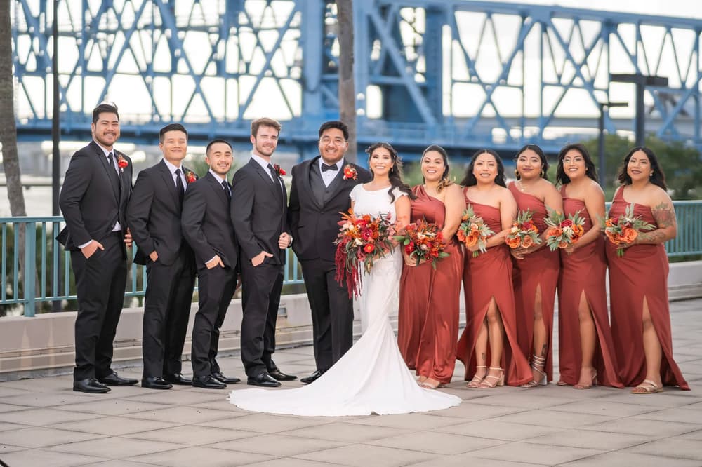 A vibrant wedding party poses together on a bridge with a striking blue steel structure in the background under golden hour lighting.