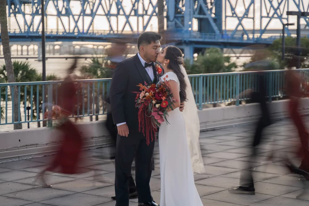 A bride and groom share a tender kiss on a bridge with blurred figures in the foreground during golden hour.