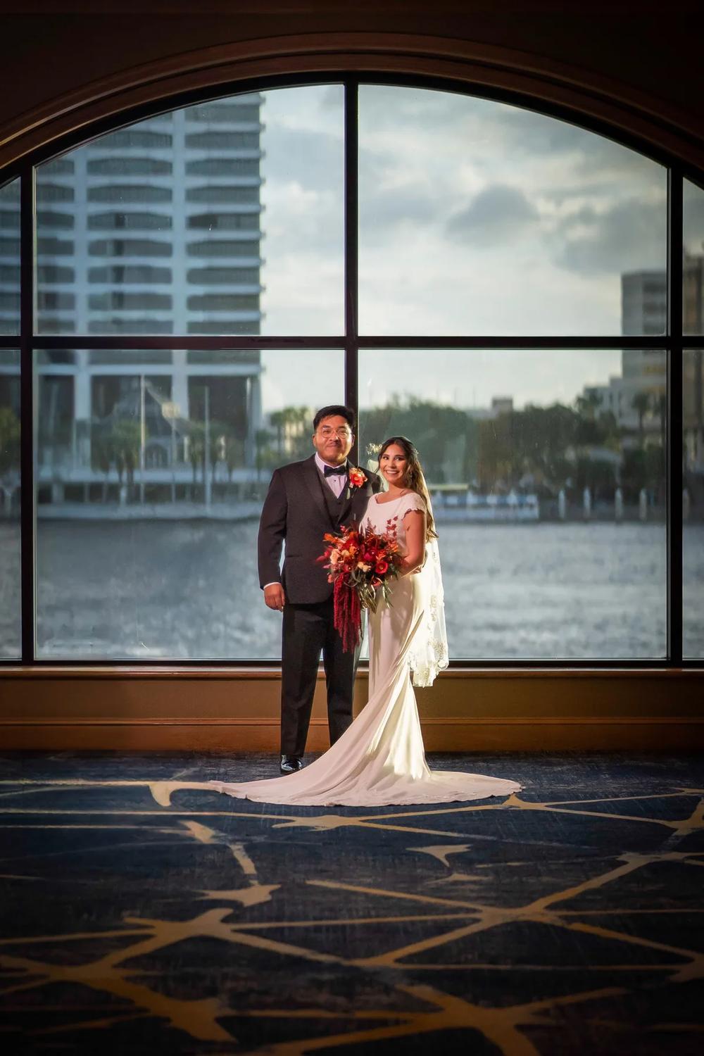 A bride and groom stand together in front of a large window with city views, bathed in golden hour light, exuding a romantic ambiance.
