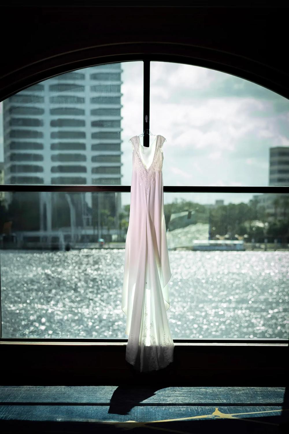 A delicate wedding gown hangs gracefully against a window overlooking a sparkling waterfront and modern cityscape under soft natural light.