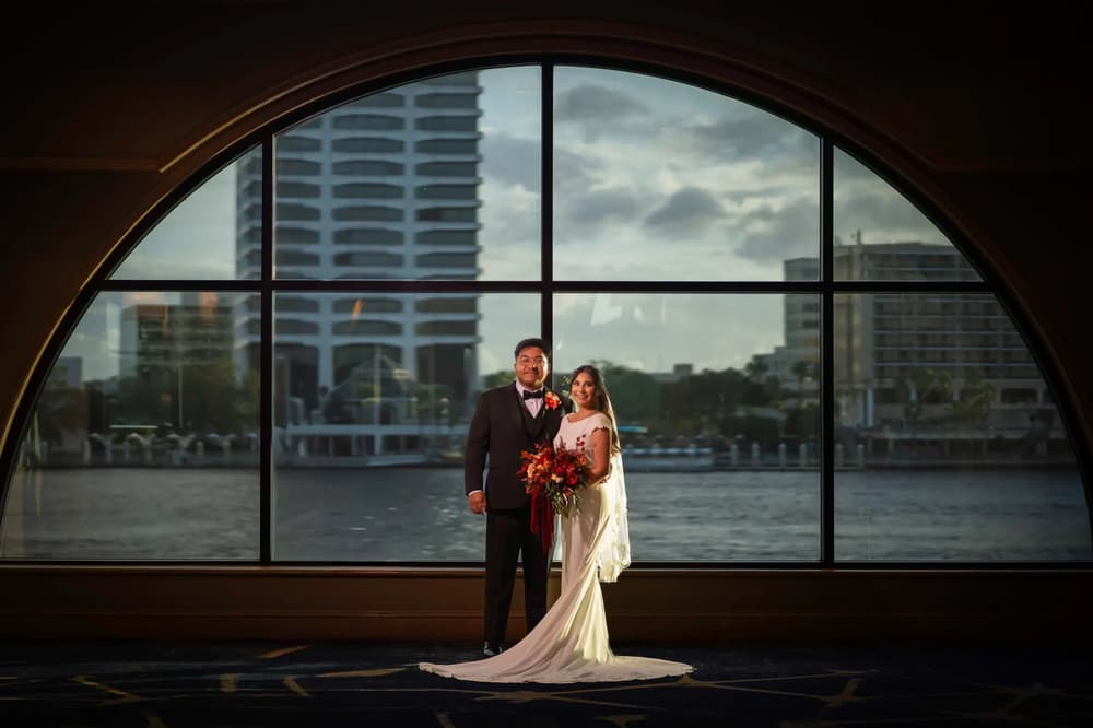 A bride and groom stand together in front of a large arched window with a cityscape view during the golden hour.