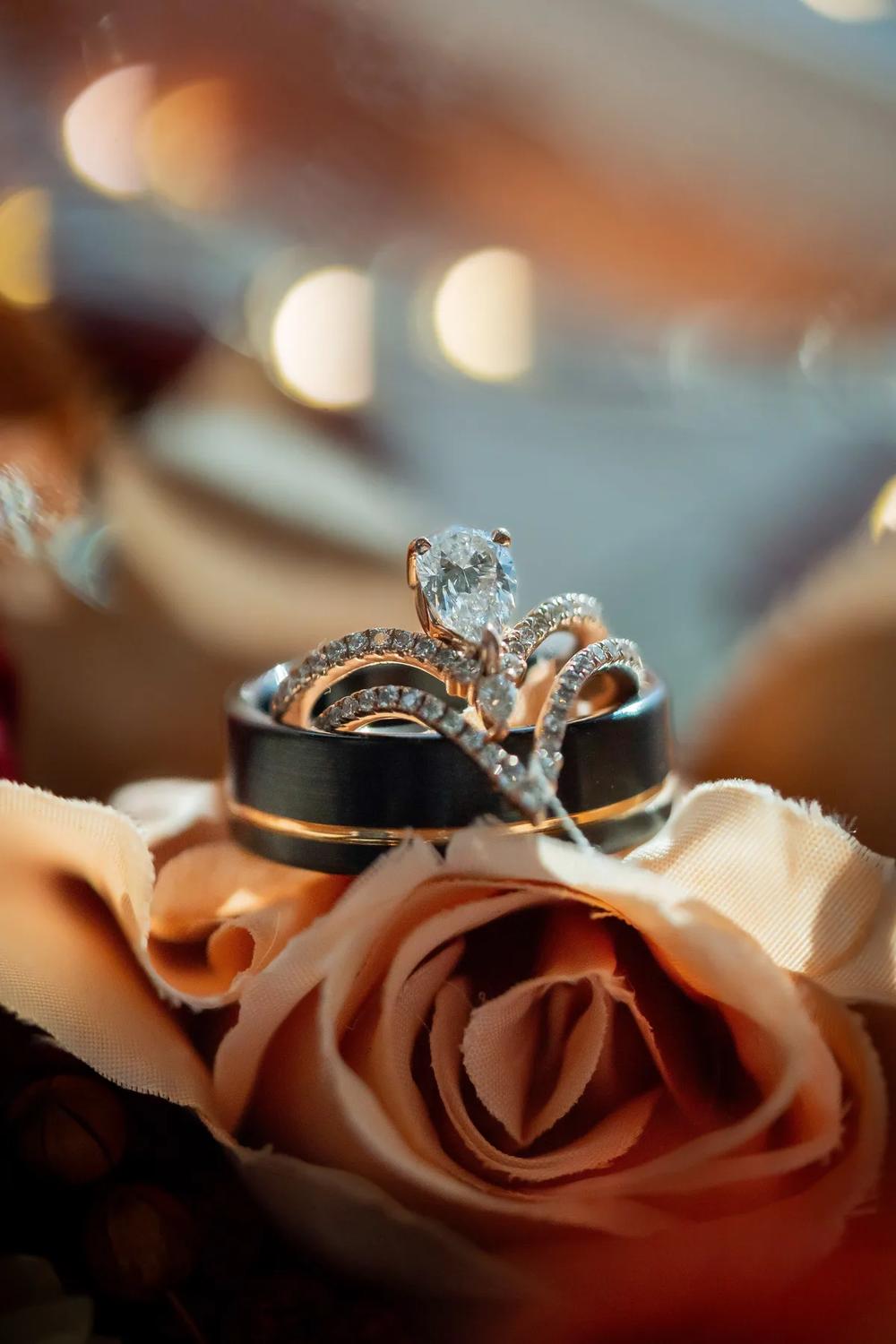 A close-up of a wedding ring set resting on a delicate rose against a softly blurred bokeh background.