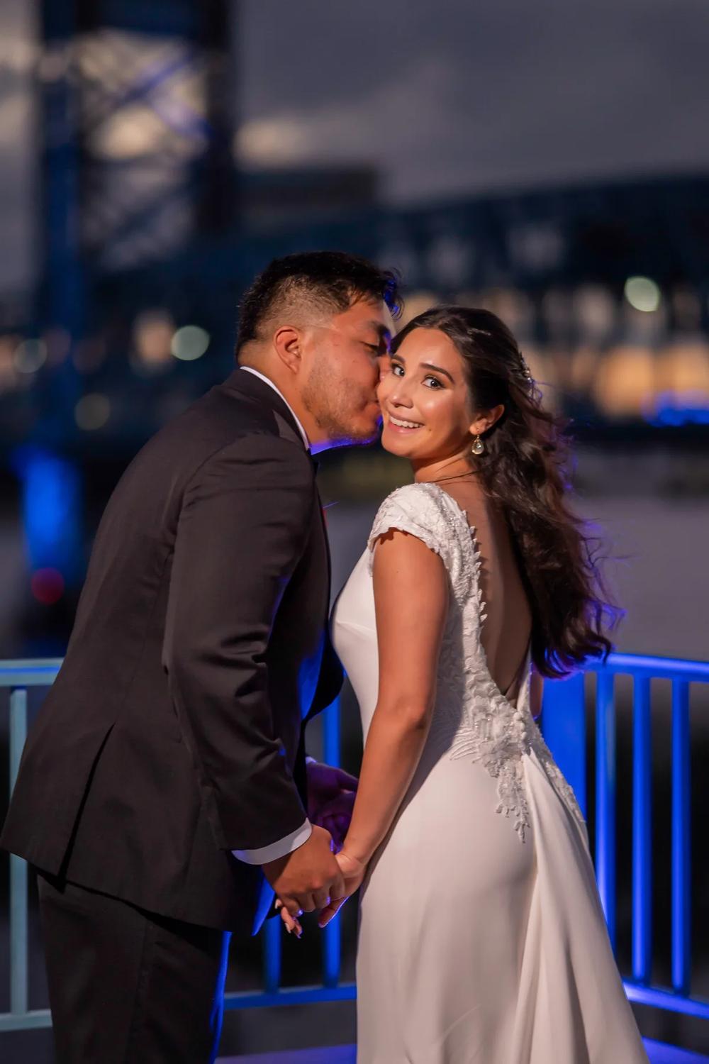 A bride and groom share a tender moment against the backdrop of a city skyline at night.