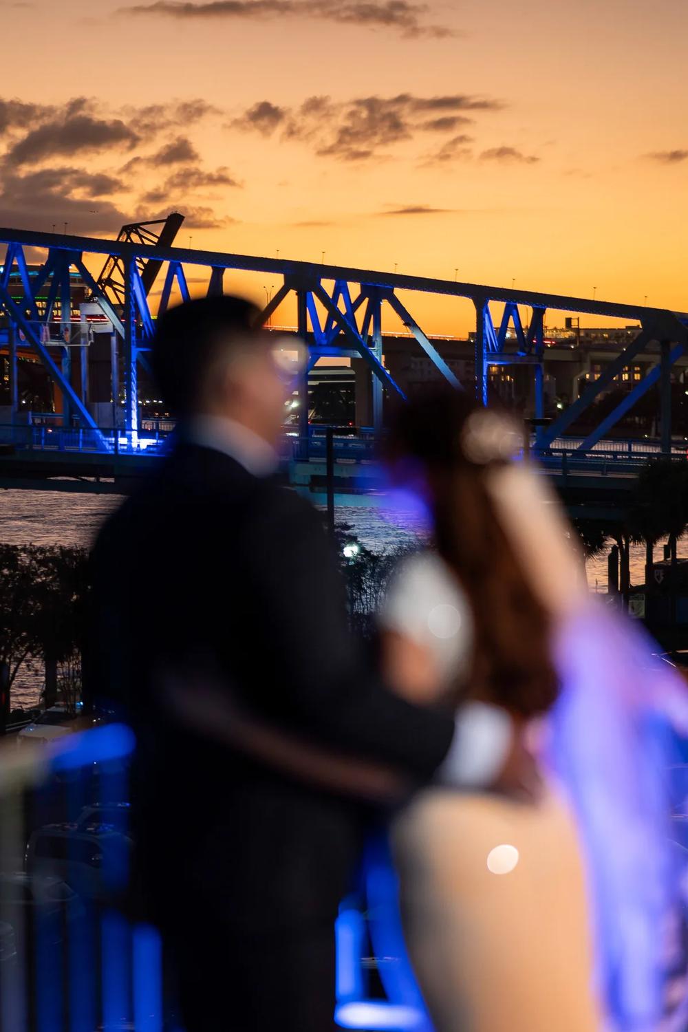 A romantic couple embraces against the backdrop of a vibrant sunset and illuminated bridge.