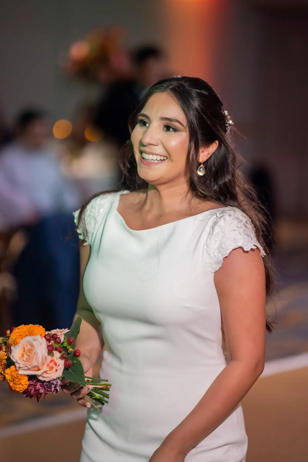 A radiant bride smiles brightly while holding a bouquet of vibrant flowers in an indoor setting with warm lighting.