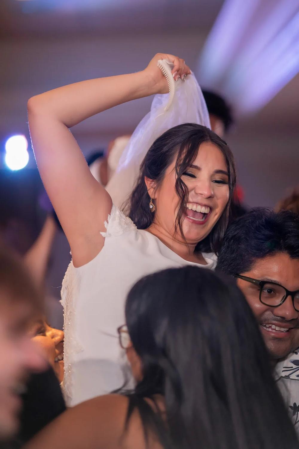 A bride joyfully dances amidst a crowd at an evening wedding reception under vibrant stage lighting.