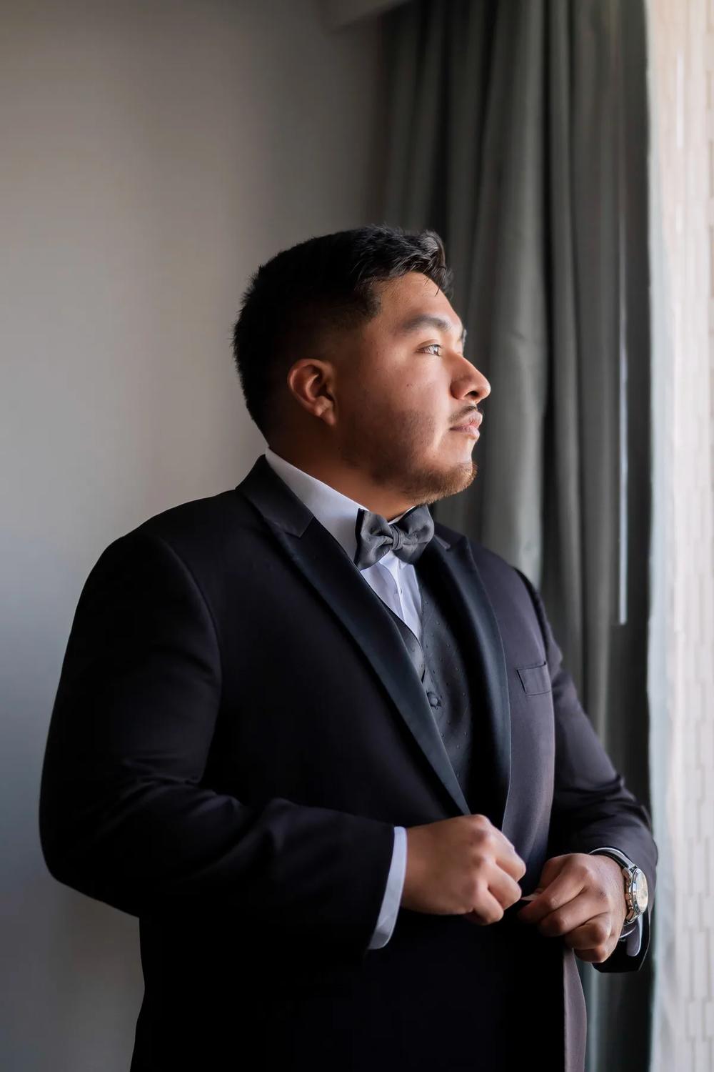 A groom in a black tuxedo stands by the window, adjusting his jacket with a contemplative expression.