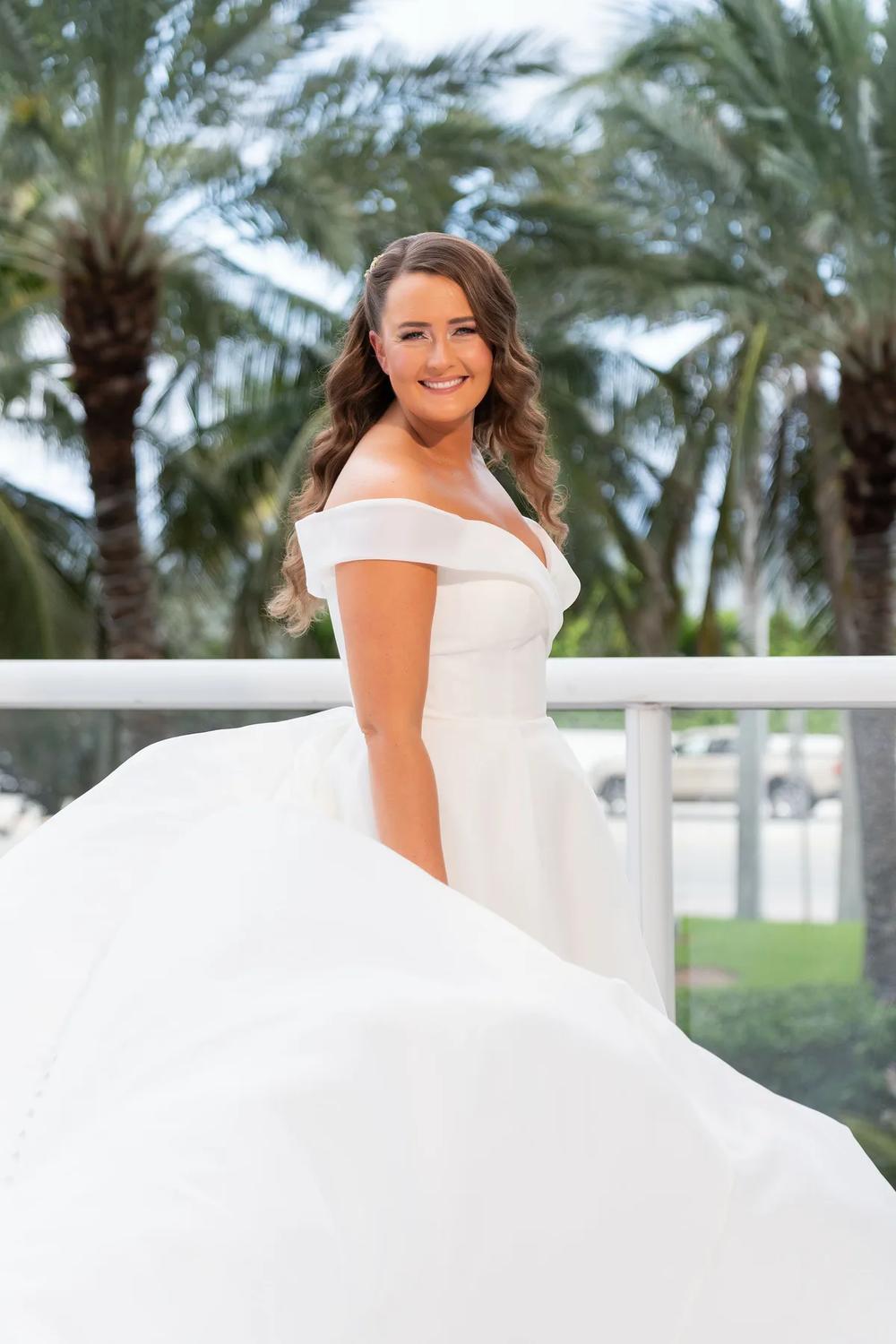 A bride in a flowing white gown smiles brightly against the backdrop of palm trees on a sunny day.