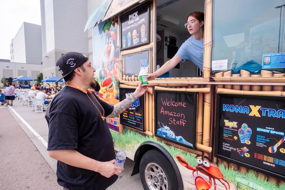 A vibrant scene at an Amazon staff event with a Kona Ice truck serving refreshing treats under bright daylight.