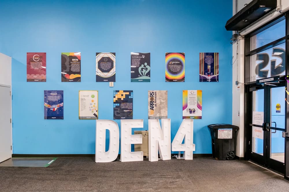 A vibrant office wall display featuring Amazon employee groups and the word 'DEN4' in large white letters.
