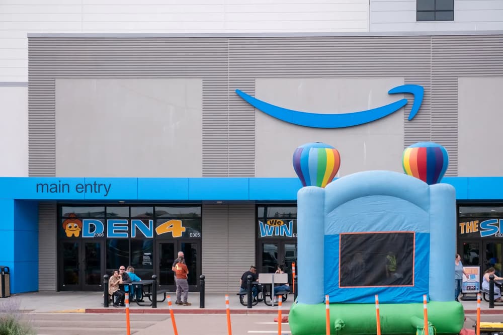 A lively outdoor scene featuring an Amazon storefront with a bouncy castle and colorful balloons, set against the backdrop of a bright blue sky.