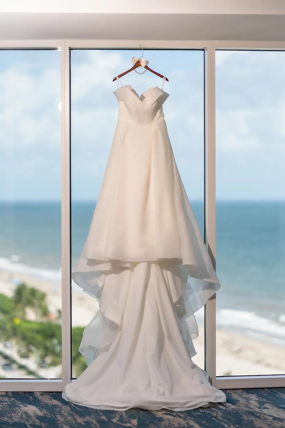 A pristine wedding gown hangs elegantly against a window overlooking the serene ocean, evoking a sense of tranquility and anticipation.