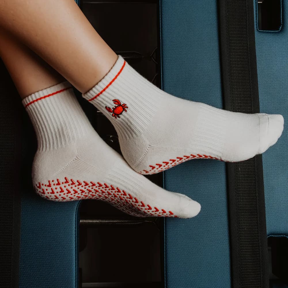 a close-up of feet wearing white socks with a red crab and textured soles, resting on the edge of an open suitcase.