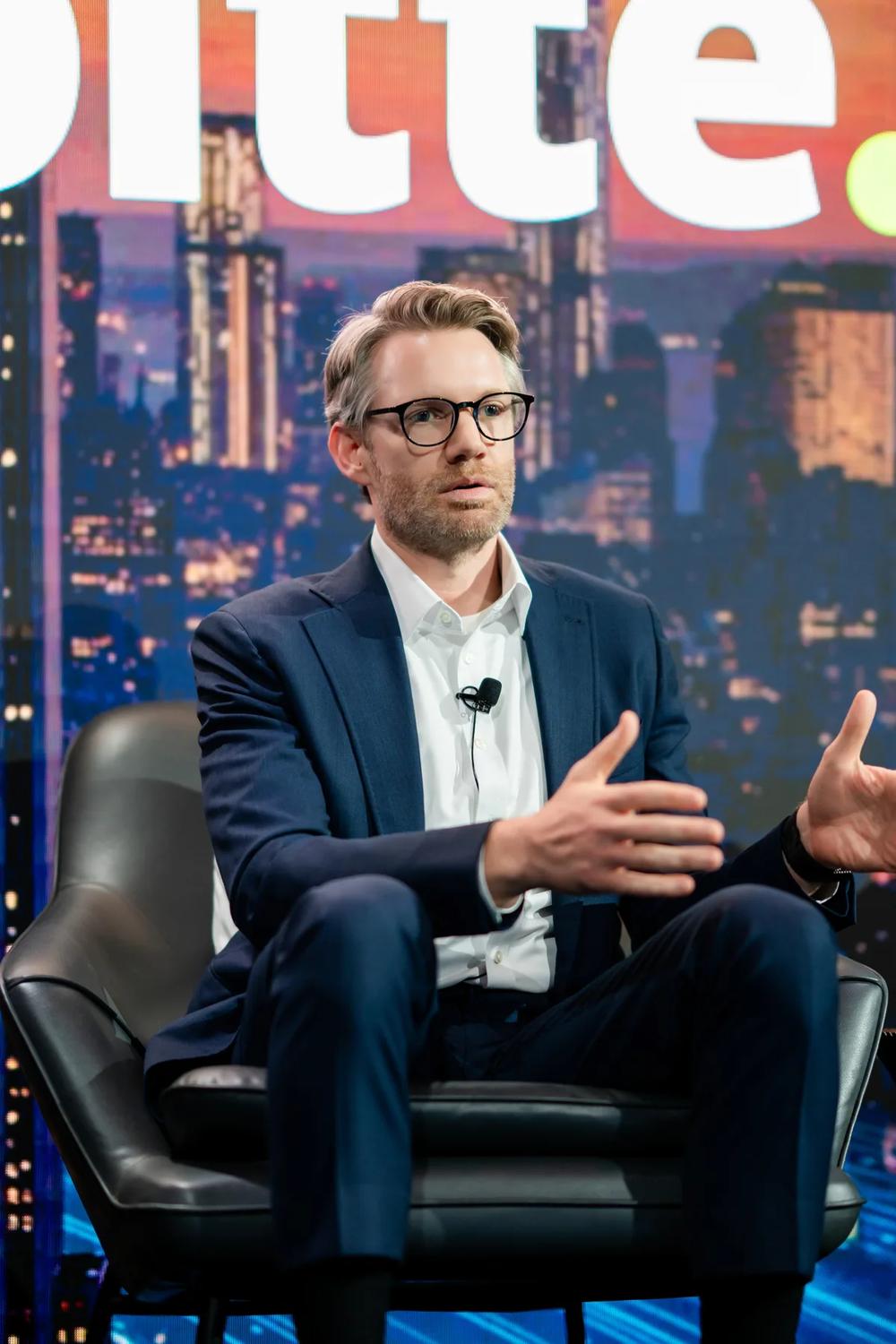 a man in a suit is seated and gesturing during an interview on a talk show set with a cityscape backdrop.