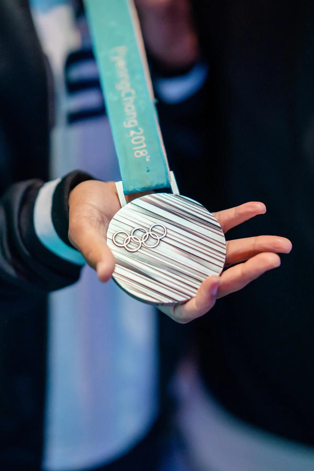 a hand holding a PyeongChang 2018 Olympic medal with the Olympic rings prominently displayed.