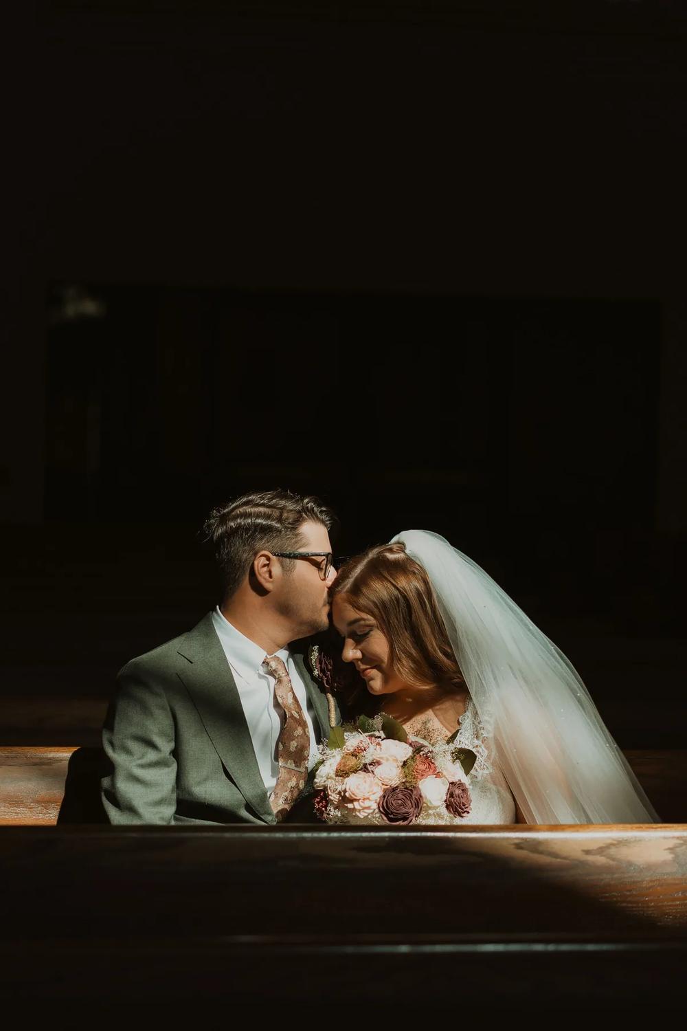 A tender moment between a bride and groom in a dimly lit church pew, bathed in soft golden light.