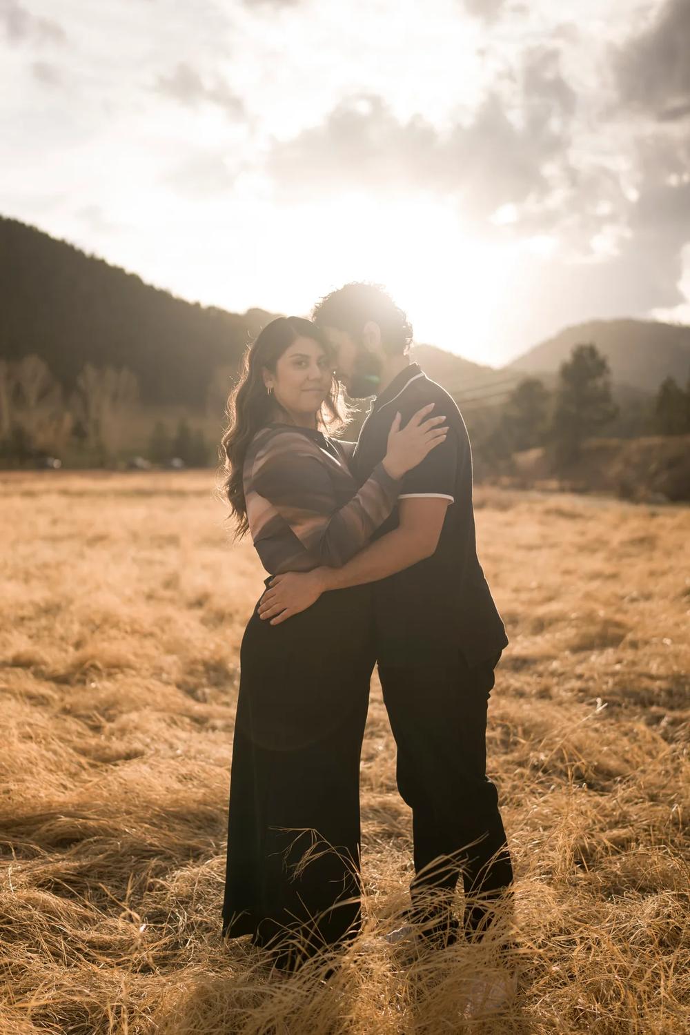 A man and a woman are standing together with their arms around each other. They are both smiling at the camera. The sun is shining on them creating a silhouette effect. The background consists of grass and mountains.