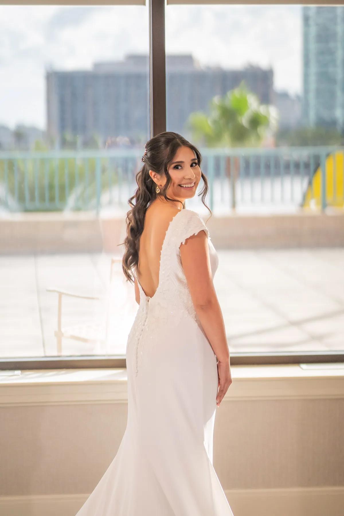 A bride in a stunning white gown smiles over her shoulder against a bright window backdrop with city views.
