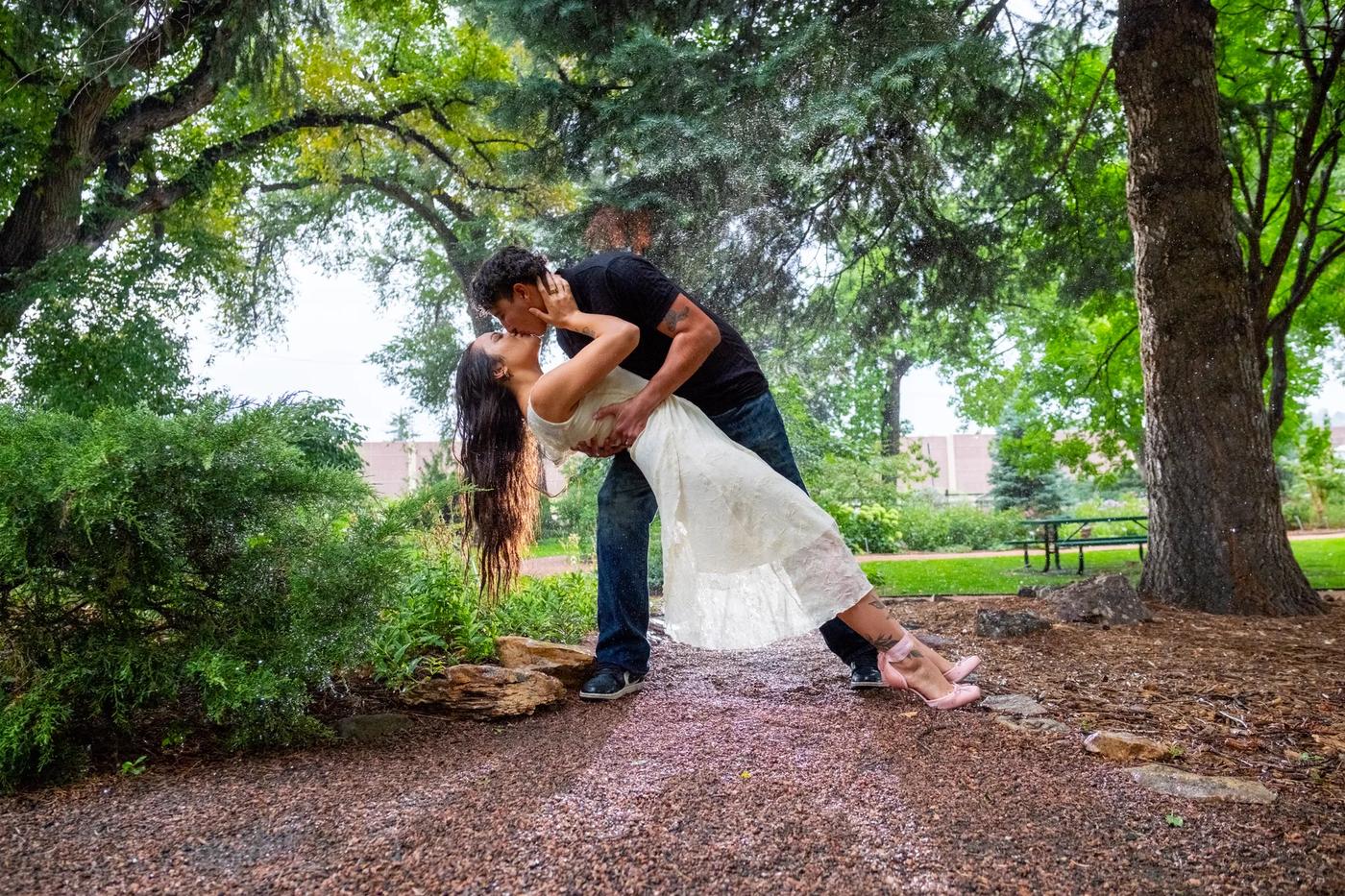 A couple shares an intimate kiss amidst a shower of rain in a lush outdoor garden setting.