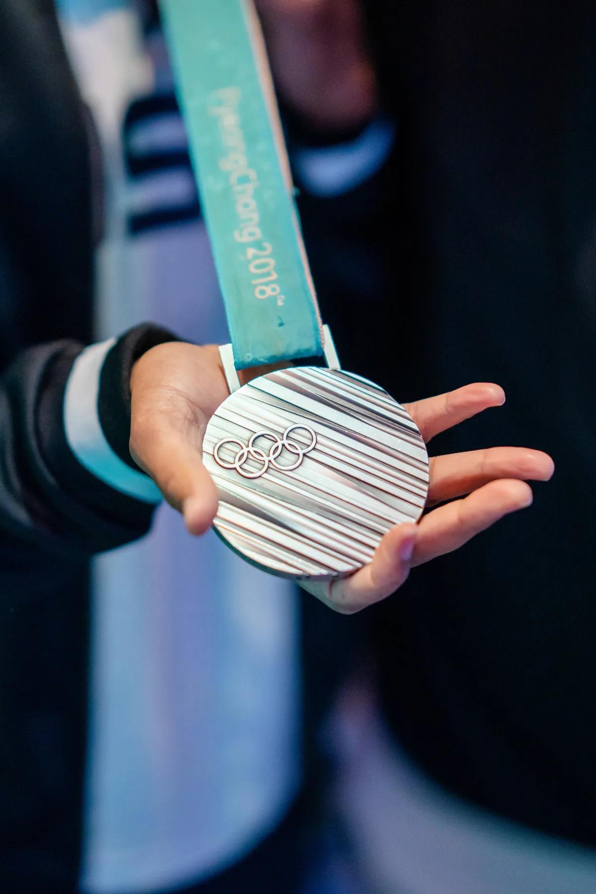 a hand holding a PyeongChang 2018 Olympic medal with the Olympic rings prominently displayed.