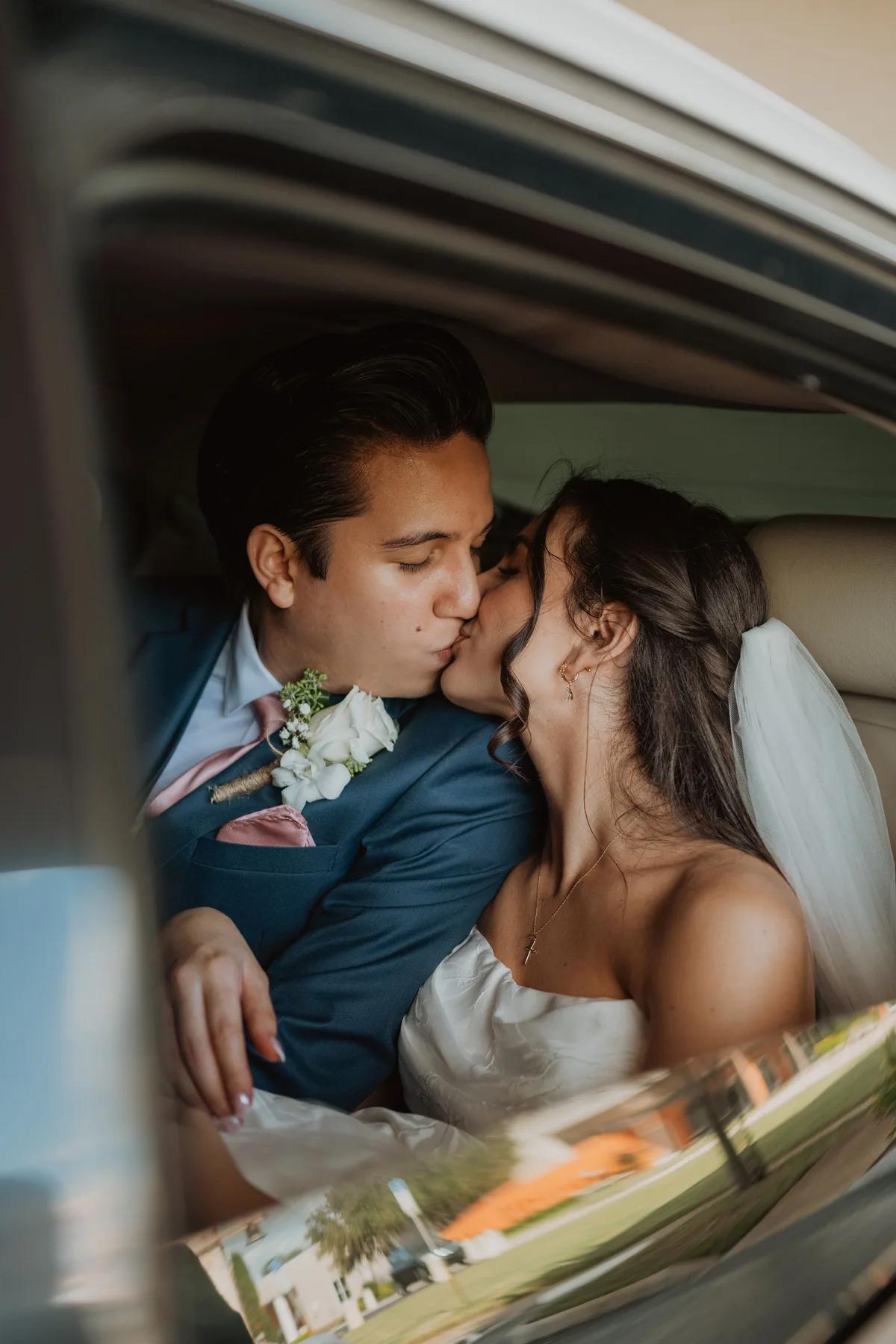 A man wearing a blue suit with white flowers on his lapel leans over to kiss a woman who is wearing a white dress. They are sitting in the back of a car together. The background shows an outdoor scene with trees and grass.