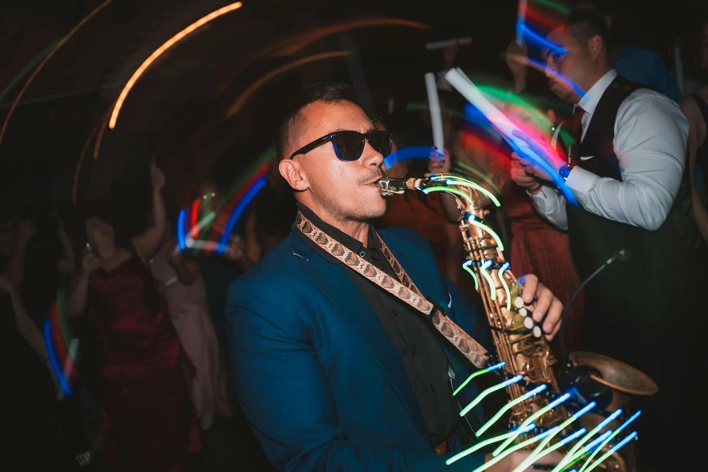 A saxophonist in a vibrant blue suit and sunglasses performs passionately at an evening wedding reception with neon lights swirling around him.