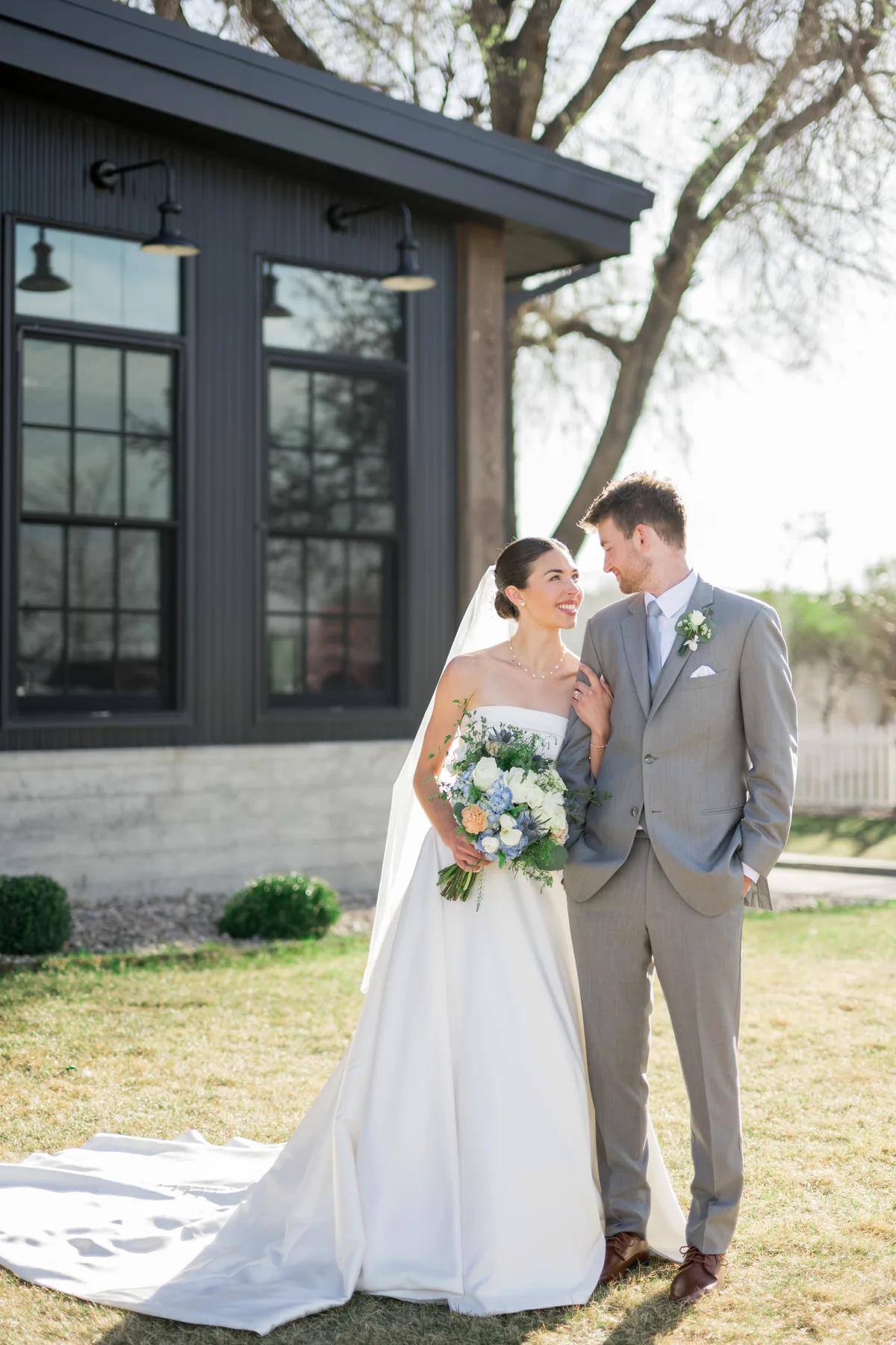 * A bride and groom standing together outside of a building. The man is wearing a gray suit with white flowers on his lapel. The woman is wearing a long white dress with greenery in her hair. * They are holding hands and smiling at each other. * Behind them, there is a black building with windows and lights hanging from the ceiling