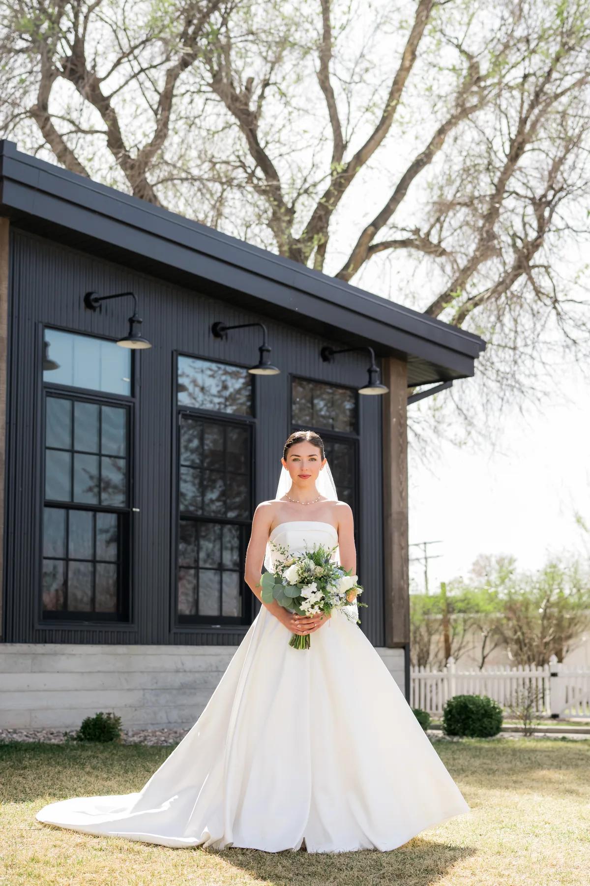 A bride holds a bouquet of flowers while standing outside her house.