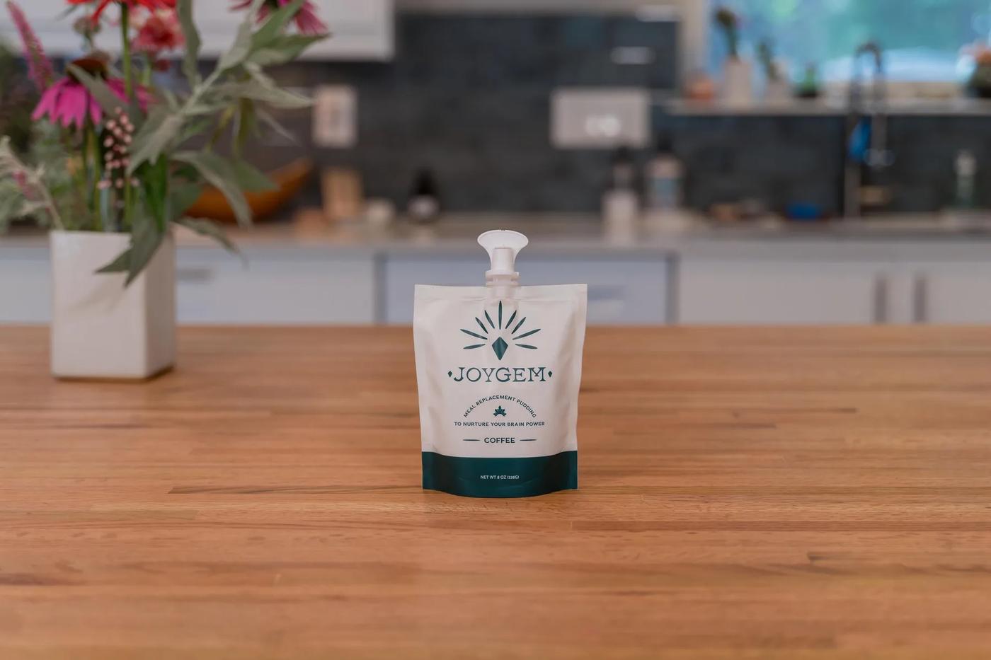 A JOYGEM meal replacement pudding pouch sits on a wooden countertop in a modern kitchen setting with soft natural light and fresh flowers in the background, creating a calm and inviting mood.