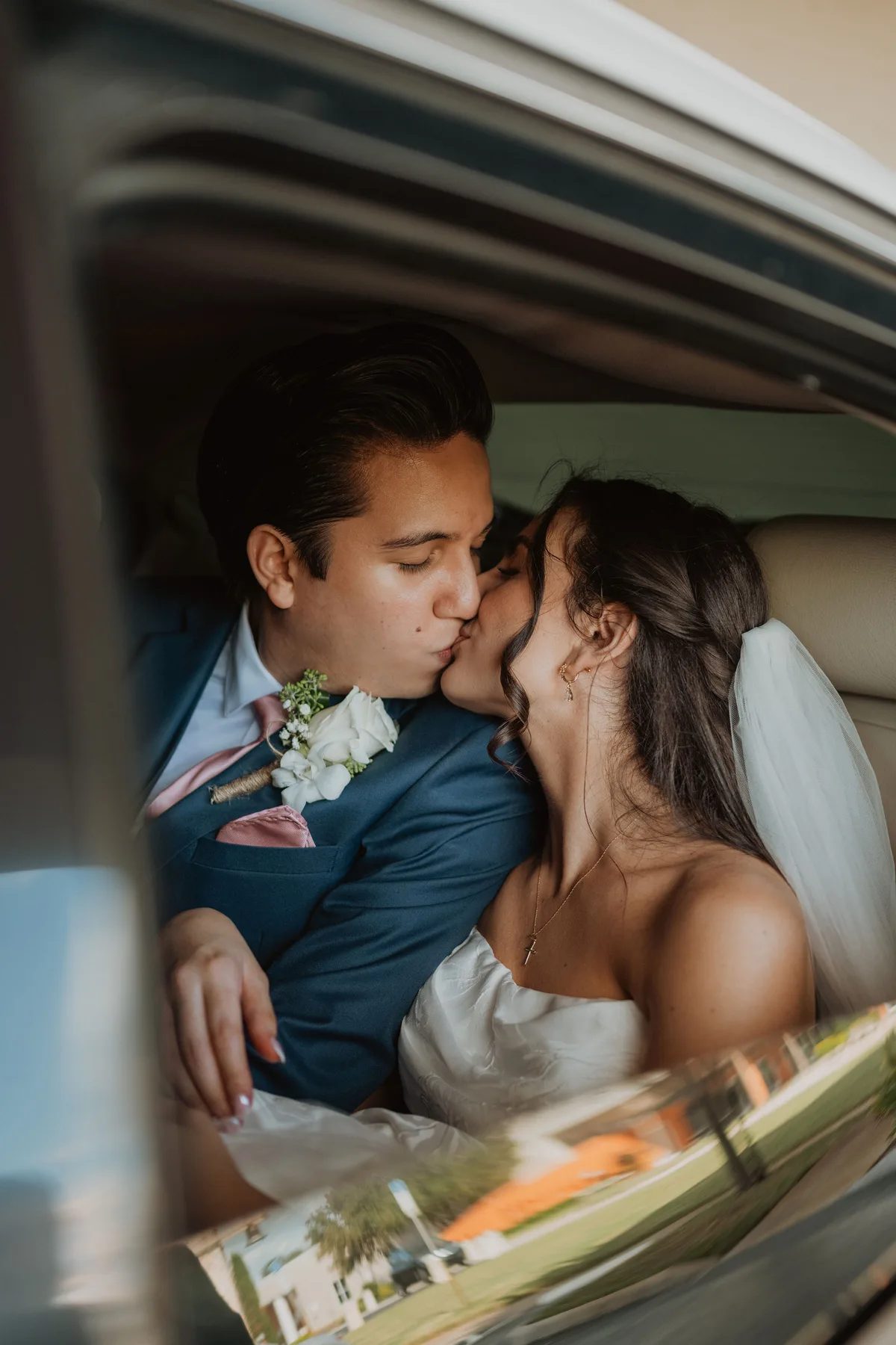 A man wearing a blue suit with white flowers on his lapel leans over to kiss a woman who is wearing a white dress. They are sitting in the back of a car together. The background shows an outdoor scene with trees and grass.