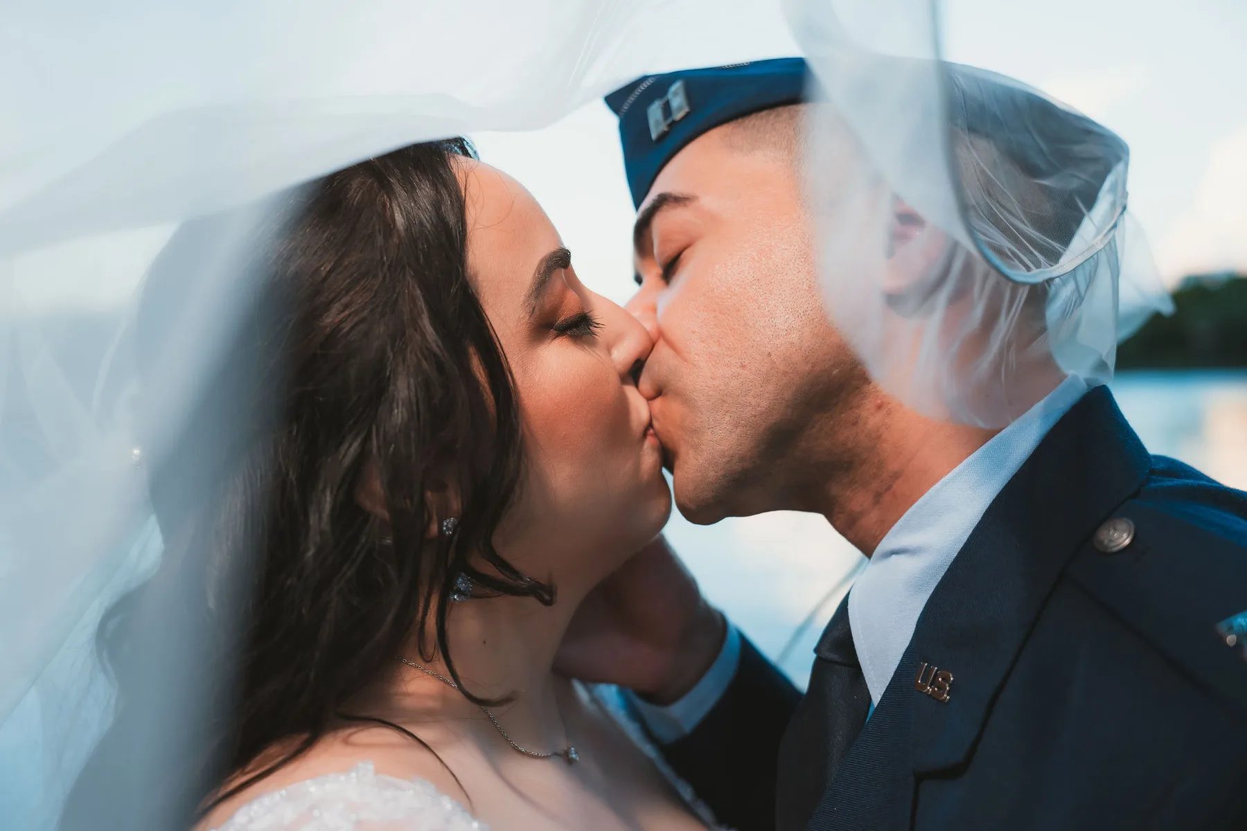 A bride and groom share a tender kiss beneath their veil against the backdrop of an outdoor setting during golden hour.