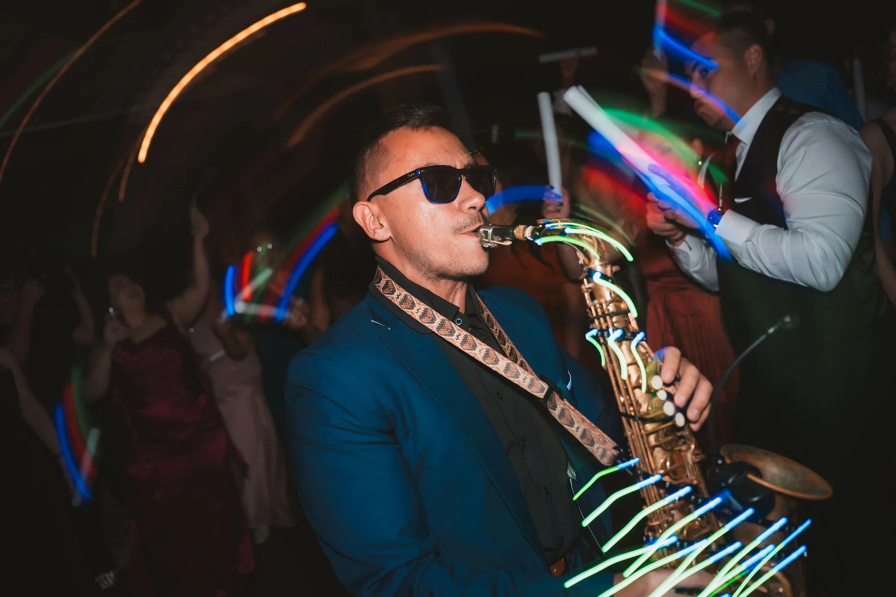A saxophonist in a vibrant blue suit and sunglasses performs passionately at an evening wedding reception with neon lights swirling around him.