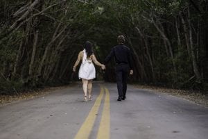 A man and a woman walking hand in hand on a road with trees around them.