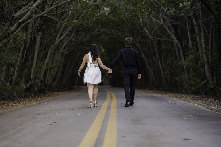 A man and a woman walking hand in hand on a road with trees around them.