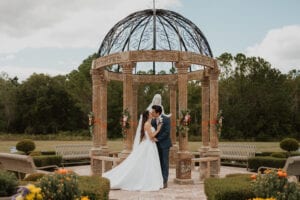 A man and a woman are standing under an archway with flowers on it. The man is wearing a blue suit and the woman is wearing a white dress. They are both looking at each other and smiling. The setting is outdoors in a garden with trees, bushes, and benches around them. The mood of the image is happy and romantic.