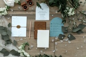 A wooden table with a bunch of wedding stationery on it including an invitation card, a guest list, place cards, a menu and a seating plan. There are also some white flowers scattered around the table. The setting is outdoors in springtime giving off a romantic mood.